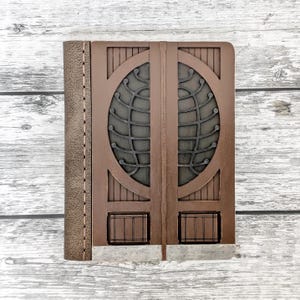 Puede incluir: Un diario de cuero marrón con una cubierta frontal decorativa con un diseño de globo terráqueo. La cubierta tiene un patrón geométrico marrón oscuro con un lomo marrón claro y un borde cosido. El diario descansa sobre una superficie de madera clara.