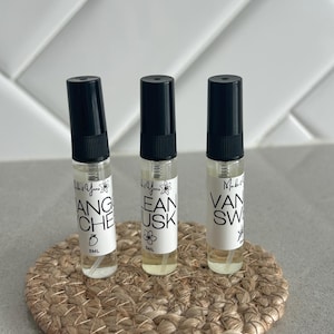 May include: Three small perfume bottles with black lids and white labels, each with a different scent name. The bottles are on a round, woven coaster. The labels read "Mango", "Clean Musk", and "Vanilla Sweet".