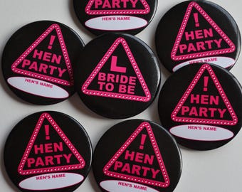 May include: A collection of black round buttons with a pink triangle design. The triangle contains the words "HEN PARTY" and an exclamation point. One button reads "BRIDE TO BE". Each button has a space for a name.