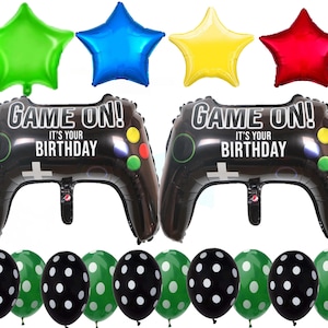 Op de afbeelding: Een set feestversiering voor een verjaardagsfeest met een videogamethema. De set bevat twee grote, zwarte en groene opblaasbare videogamecontrollers met de tekst "Game On! It's Your Birthday", vier kleurrijke stervormige ballonnen en acht stippenballonnen in zwart en groen.