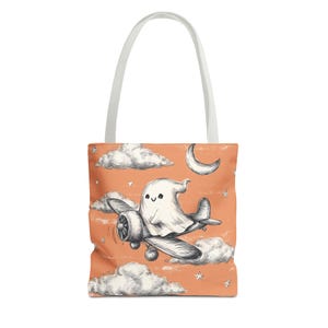 May include: Tote bag with a whimsical illustration of a ghost flying a biplane against a peach-colored sky with clouds, a crescent moon, and stars. The bag has a white handle.