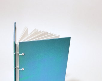 Può includere: Un quaderno blu turchese con una rilegatura bianca e anelli argentati. Il quaderno è aperto e le pagine sono a ventaglio.