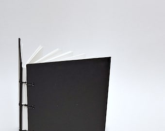 May include: A black leather-bound journal with white pages and black stitching. The journal is closed and standing upright on a white surface.