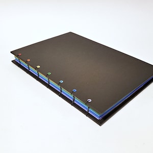 May include: A black notebook with a rainbow of colored pages bound together with black thread and colorful plastic rings.