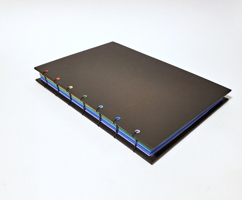 May include: A black notebook with a rainbow of colored pages bound together with black thread and colorful plastic rings.