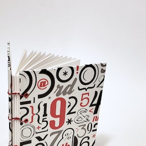 May include: A white notebook with a red and black pattern of numbers, letters, and symbols. The notebook is open and the pages are fanned out.