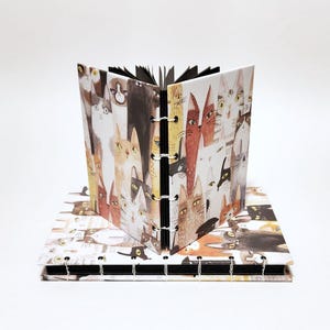 May include: A handmade open book with a cat-themed cover and pages. The cover and pages are illustrated with various cat faces in different colors and patterns. The book is bound with white thread and has black edges.