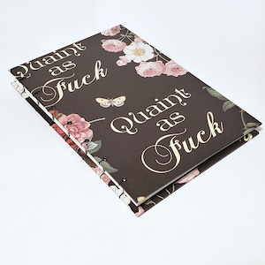 May include: A spiral-bound notebook with a dark brown cover featuring the text "Quaint as Fuck" in elegant script. The cover is adorned with pink roses, white flowers, green leaves, and a small butterfly illustration.