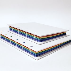 May include: Two rainbow-colored journals with white covers. The journals are stacked on top of each other. The top journal is slightly larger than the bottom journal. The journals are bound with a rainbow of colored paper.