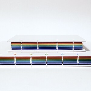 May include: Two rainbow-colored journals with white covers and white stitching. The journals are stacked on top of each other.