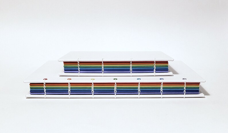 May include: Two rainbow-colored journals with white covers and white stitching. The journals are stacked on top of each other.