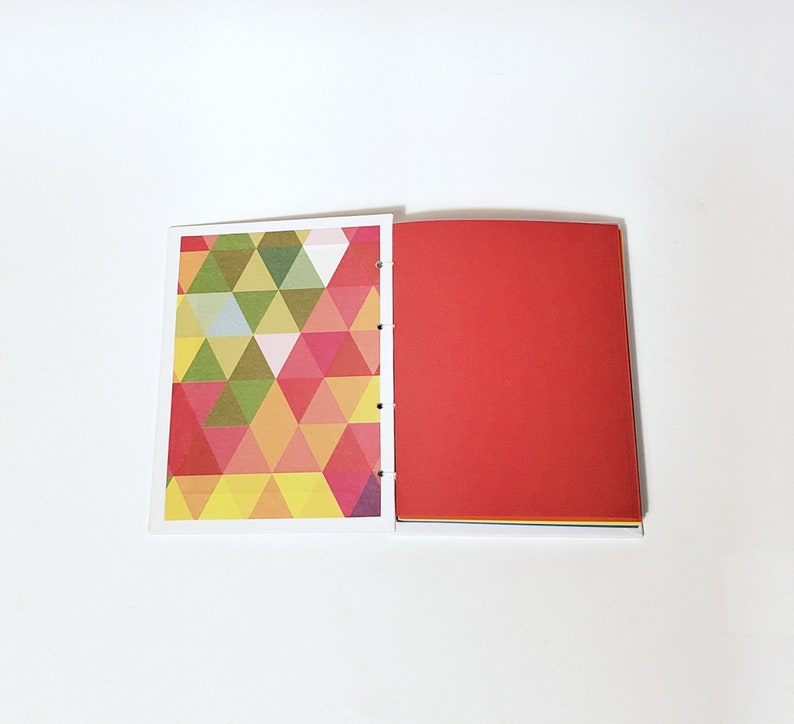 May include: A notebook with a geometric pattern on the front cover and a solid red back cover. The notebook is open to reveal blank pages.