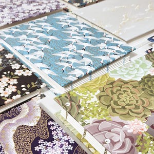 May include: A collection of handmade journals with various patterned covers. Designs include white cranes on a blue background, floral patterns, and succulent illustrations. The journals are bound with visible stitching.