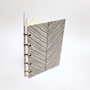 May include: A handmade journal with a black and white diagonal striped pattern on the cover. The journal is bound with black thread and has white interior pages. The journal is open, revealing the blank pages.