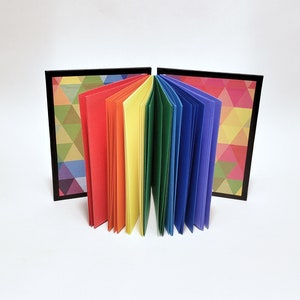 May include: A rainbow-colored notebook with a geometric pattern on the cover. The notebook is open and the pages are fanned out.