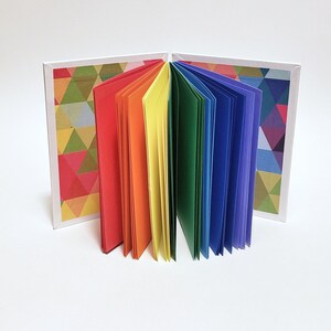 May include: A small rainbow-colored book with a geometric pattern on the cover. The book is open and the pages are fanned out.