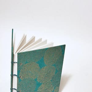 May include: A teal blue journal with a gold floral pattern on the cover. The journal is open to reveal blank white pages.