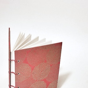 May include: A red journal with a gold floral pattern on the cover. The journal is open to reveal blank white pages.