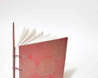 May include: A red journal with a gold floral pattern on the cover. The journal is open to reveal blank white pages.