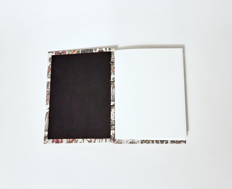 Pu&ograve; includere: Un quaderno aperto con una copertina nera e una pagina bianca. La copertina &egrave; decorata con un motivo colorato di persone e animali.