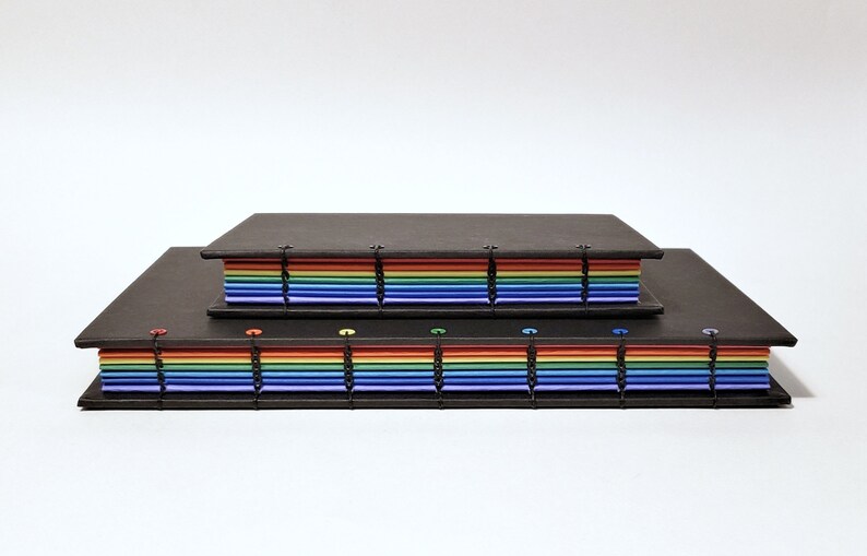May include: Two black bound journals with rainbow colored pages. The smaller journal is stacked on top of the larger journal.