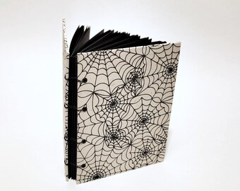 May include: A small, open book with a spiderweb design. The cover is off-white with a black spiderweb pattern. The pages are black, and the binding is a black spiral. The book is standing upright against a white background.