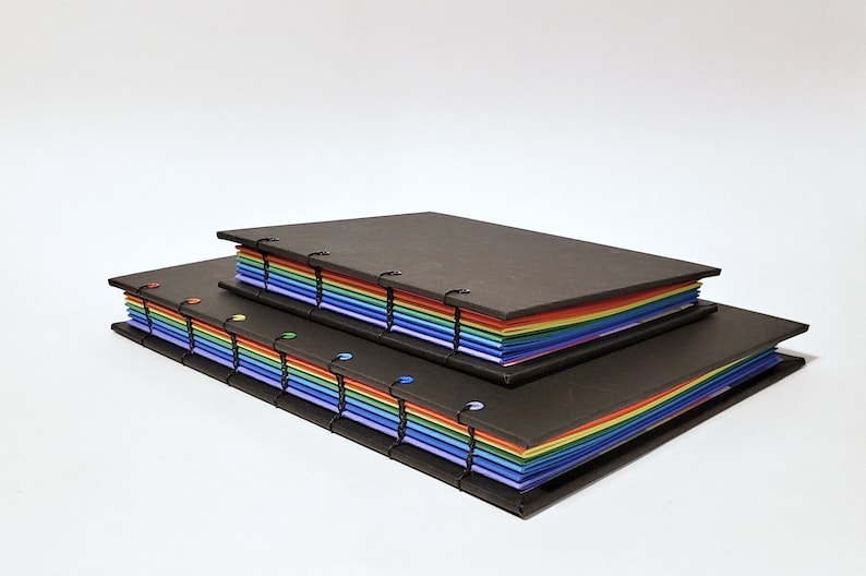 May include: Two black covered journals with rainbow colored pages. The smaller journal is stacked on top of the larger journal.