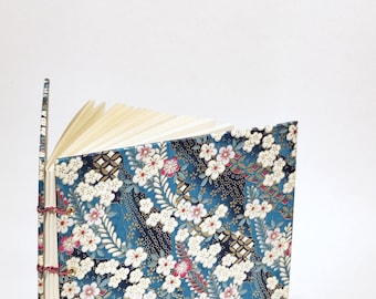 May include: A handmade journal with a floral patterned cover in shades of blue, white, and red. The journal is open, revealing the blank pages inside. The binding is visible with red thread.