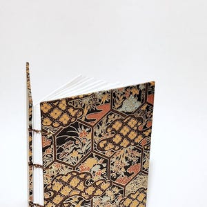 May include: A black, gold, and red patterned journal with a white spine and pages. The cover features a repeating pattern of hexagonal shapes with floral and cloud designs.