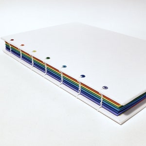 May include: A white notebook with a rainbow spine. The spine is made of colored paper and has small metal rings holding the pages together.
