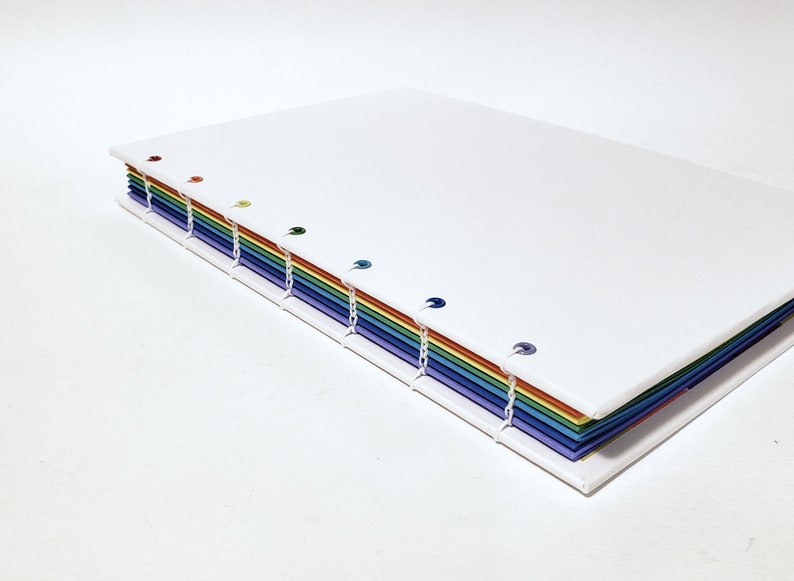 May include: A white notebook with a rainbow spine. The spine is made of colored paper and has small metal rings holding the pages together.