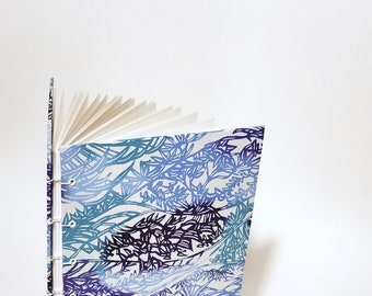 May include: A blue and white patterned journal with a white spine and pages fanned out. The cover features a repeating pattern of stylized leaves and flowers.