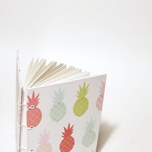 May include: A white notebook with a pineapple pattern. The pineapples are in various colors, including pink, green, and red. The notebook is bound with metal rings.