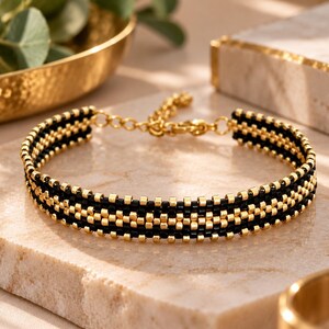 May include: A woven bracelet featuring alternating rows of small black and metallic gold beads. The design includes a gold-toned metal lobster claw clasp and an adjustable chain link extension, creating a structured and uniform beaded pattern.