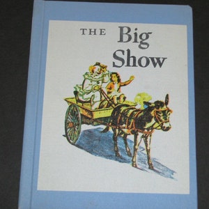 Puede incluir: Un libro azul con el título "THE Big Show" en la portada. La portada presenta una ilustración de un payaso y un niño que van en un carro tirado por un burro.