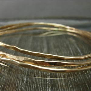 May include: Three gold-toned hammered metal bangles stacked together. The bangles are smooth and have a slightly irregular shape.