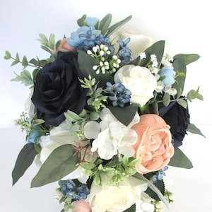 May include: A cascading bouquet featuring navy blue, white, and peach-colored roses, accented with small blue flowers and green foliage. The arrangement is designed with a variety of textures and shapes, creating a visually appealing floral display.