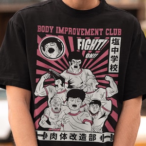 May include: Black t-shirt with a graphic design featuring the text "BODY IMPROVEMENT CLUB" and Japanese text. The design includes illustrations of muscular figures in a pink and white color scheme.