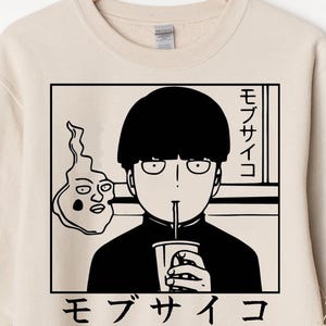 May include: Beige sweatshirt with a black and white anime graphic. The design shows a character drinking from a cup, with Japanese text and a spirit. The sweatshirt is displayed on a wooden hanger.