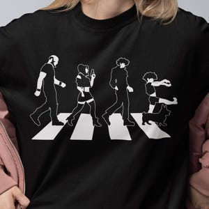Op de afbeelding: Zwart T-shirt met een witte afbeelding van vier figuren die een zebrapad oversteken, een parodie op de Abbey Road albumhoes van The Beatles. De persoon draagt een zwarte short en witte sokken.