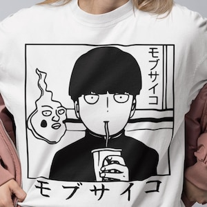 Könnte beinhalten: Weißes T-Shirt mit einer schwarz-weißen Anime-Grafik eines Charakters, der aus einer Tasse trinkt, mit japanischem Text. Die Person trägt schwarze Shorts und eine rosa Jacke. Das Bild zeigt eine sitzende Person.