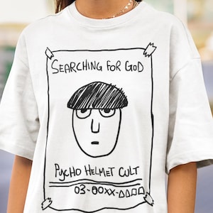 Puede incluir: Camiseta blanca con un gráfico dibujado a mano. El texto "Searching for God" está encima de una cara de dibujos animados, con "Psycho Helmet Cult" debajo. La camiseta tiene mangas cortas y cuello redondo.