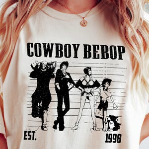 Pode incluir: Camiseta creme com um gráfico em preto e branco de personagens da série de anime Cowboy Bebop. O design inclui o texto "COWBOY BEBOP" acima dos personagens, com "EST. 1998" abaixo.