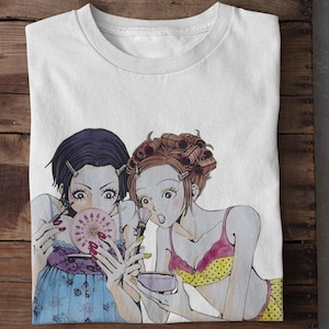 May include: A white t-shirt featuring a graphic illustration of two characters applying makeup. One character has dark hair and a blue floral top, while the other has reddish hair in curlers and a yellow polka-dot top with a pink trim.
