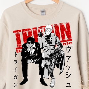 May include: Beige sweatshirt featuring a red graphic with the text "TRIGUN" and Japanese characters. The graphic depicts two anime characters in black and white. A casual, long-sleeved top.
