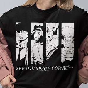 Pode incluir: Camiseta preta com um gráfico branco de quatro personagens em estilo anime em painéis verticais. O texto "SEE YOU SPACE COWBOY!" está impresso abaixo do gráfico. A pessoa está usando uma jaqueta rosa e shorts pretos.