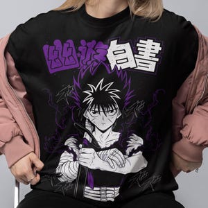 Peut inclure: Un t-shirt noir à col rond avec une impression graphique d'un personnage d'anime aux cheveux en pointe, aux bras bandés et tenant une épée. Au-dessus du personnage, le texte japonais 幽遊白書 est imprimé en blanc et violet sous une veste rose.