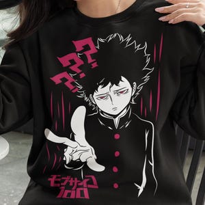 Puede incluir: Sudadera negra con un gráfico de un personaje de anime, signos de interrogación y texto japonés en blanco y rosa. La sudadera la lleva una persona que sostiene una taza negra.