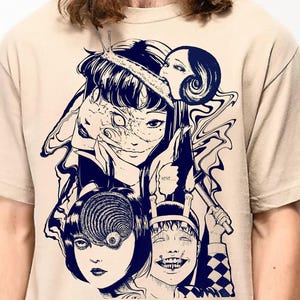May include: A beige t-shirt with a dark blue graphic print of several stylized faces with exaggerated features. The design includes a spiral eye, a smiling face with a checkered pattern, and other surreal elements. The shirt is a short-sleeved crew neck.