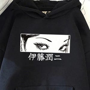 May include: Black hoodie with a white rectangular graphic displaying stylized eyes and Japanese text. The hoodie has a front pocket and a drawstring hood. The background includes a paper bag and a magazine.